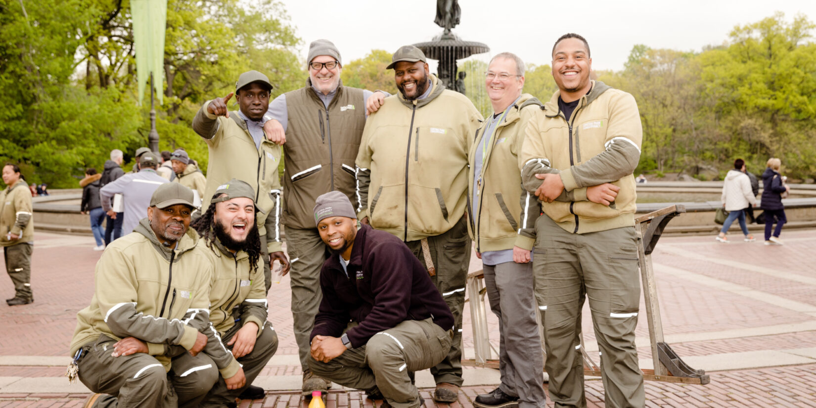 A group of Central Park Conservancy staff member in Conservancy uniform laugh and pose for the camera with Bethesda Fountain in the background.