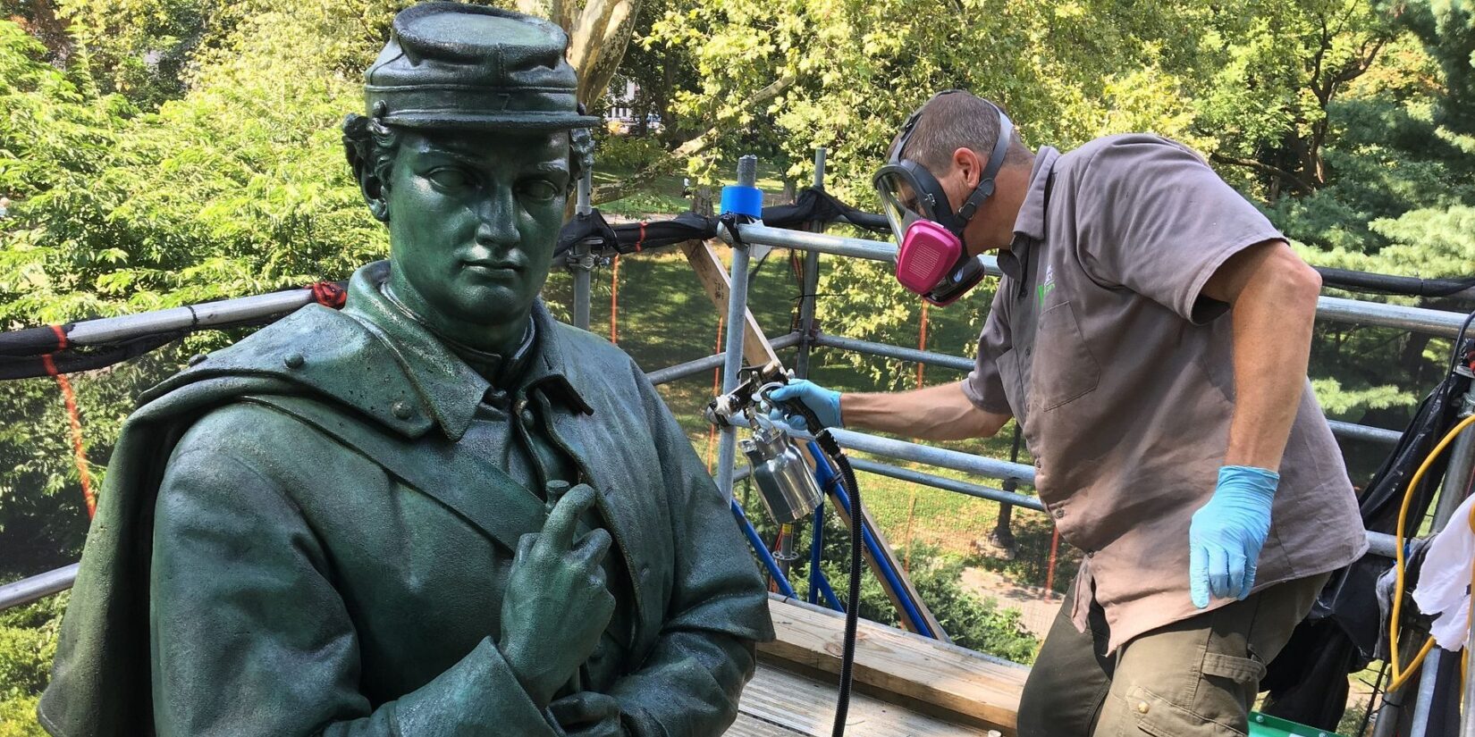 A protective coating is sprayed on the bronze statue by a worker on a scaffold.