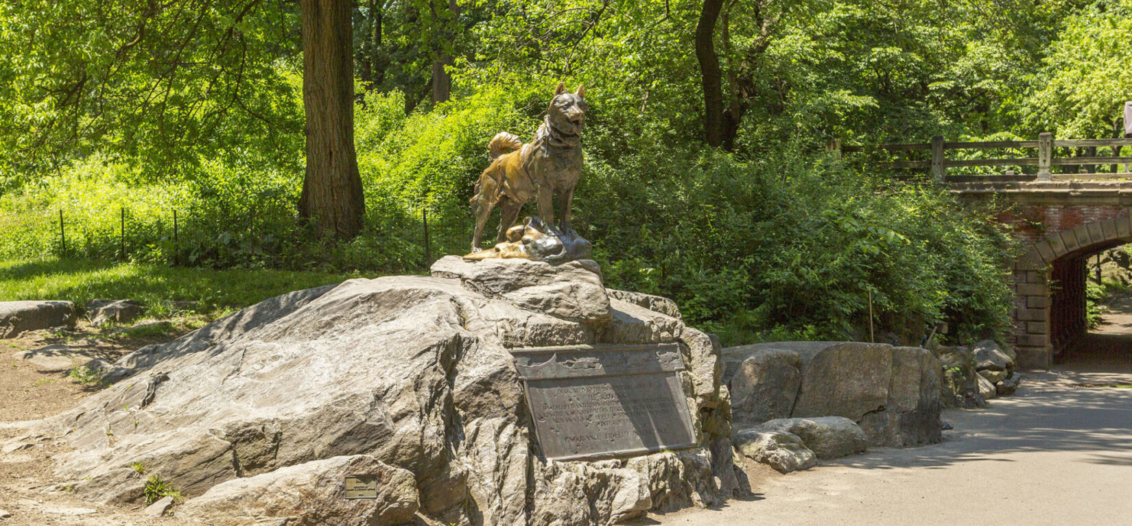 Balto is seen standing proudly on his rock, above the accompanying plaque, on a sunny day