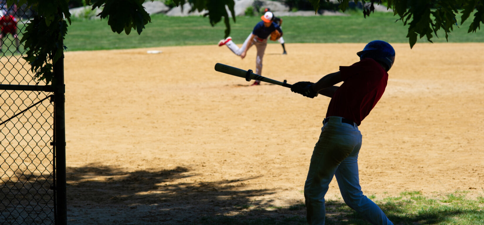 A younger player is on-deck, in the shade, ready for his at-bat