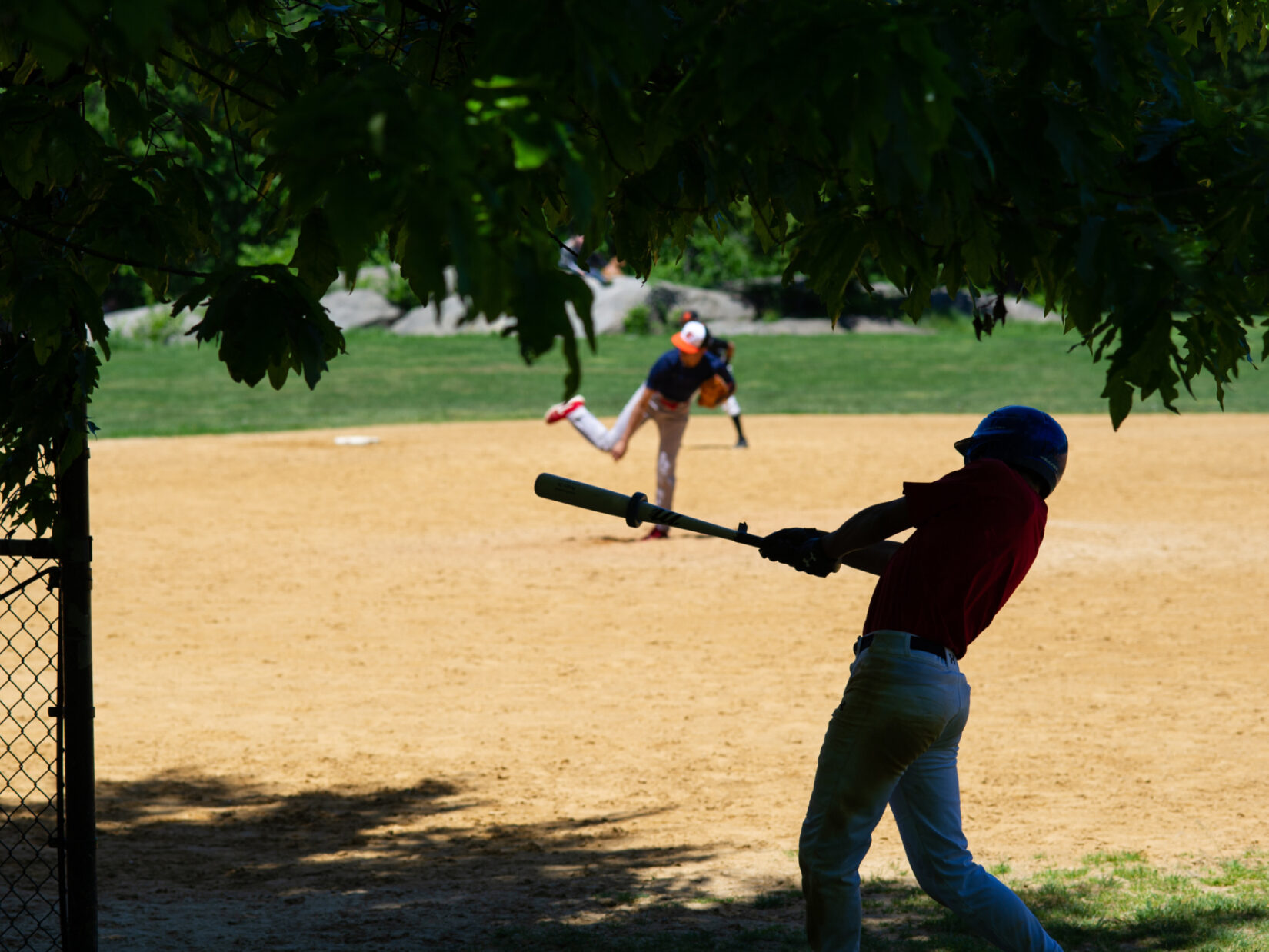 A younger player is on-deck, in the shade, ready for his at-bat