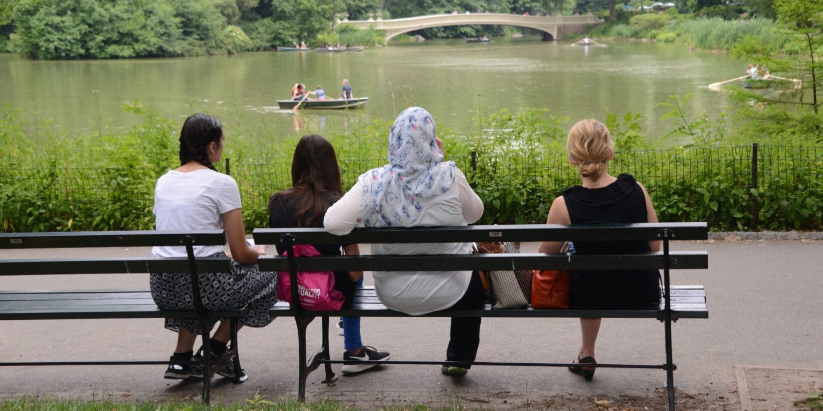 Four women, with backs to the camera, look out across the Lake to Bow Bridge in the distance.