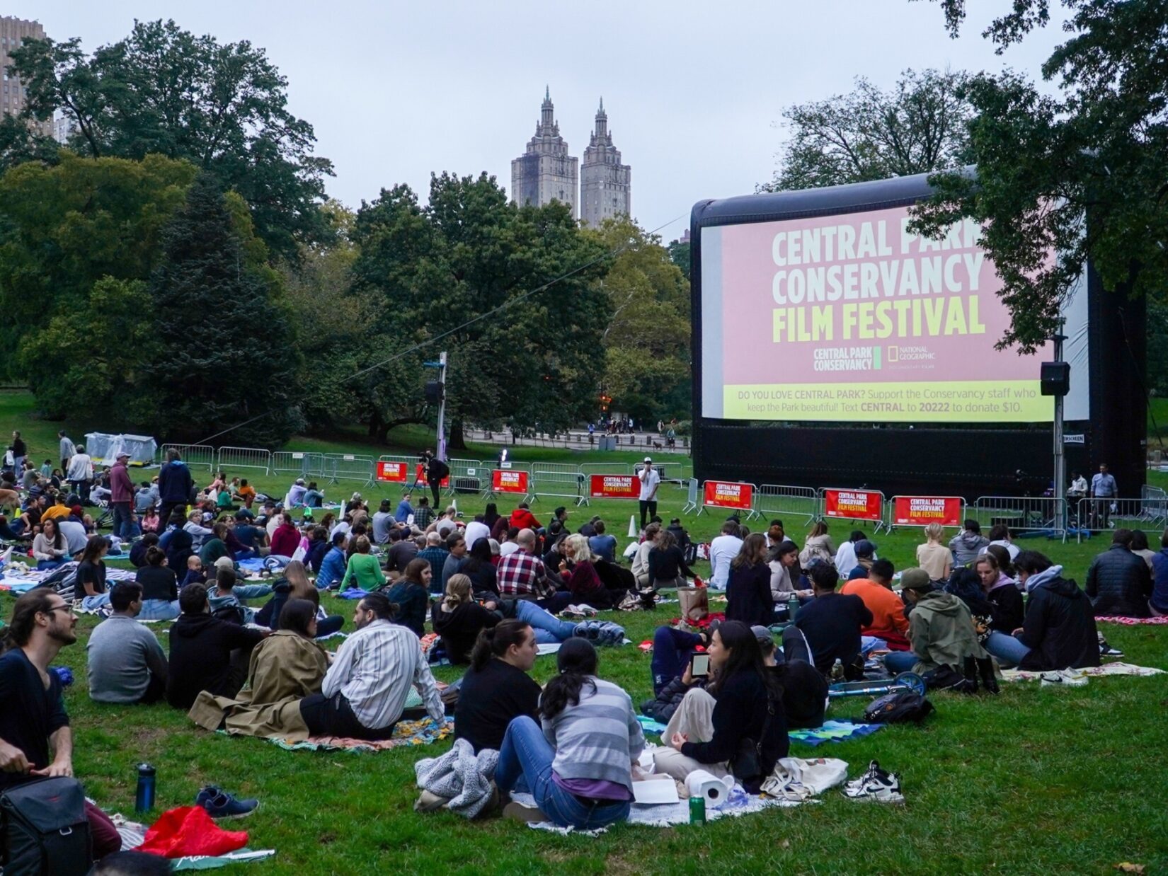 Hundreds of movie fans sit before a crowd-sized movie screen in the Sheep Medow at dusk.