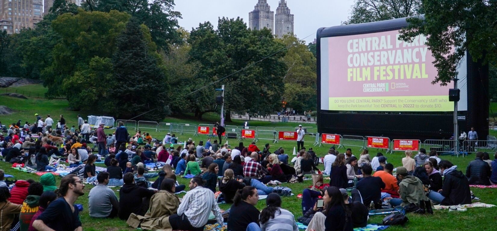 Hundreds of movie fans sit before a crowd-sized movie screen in the Sheep Medow at dusk.