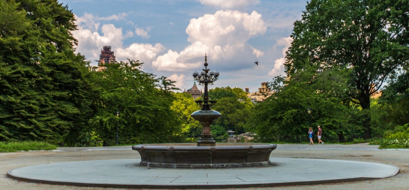 The fountain surrounded by a circular pathway lined with trees, under a blue sky with billowing clouds.