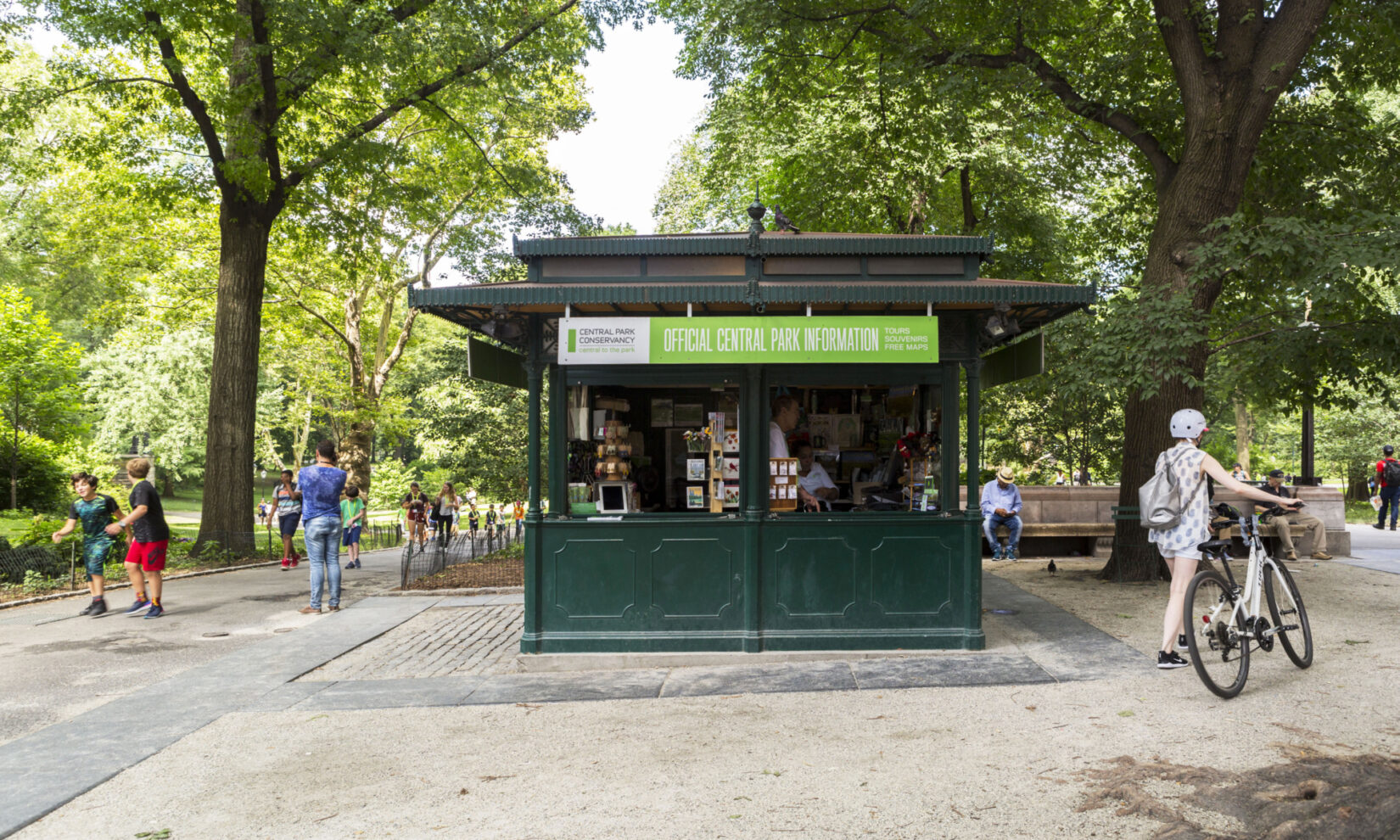 The kiosk is seen framed by summer trees and passing pedestrians.