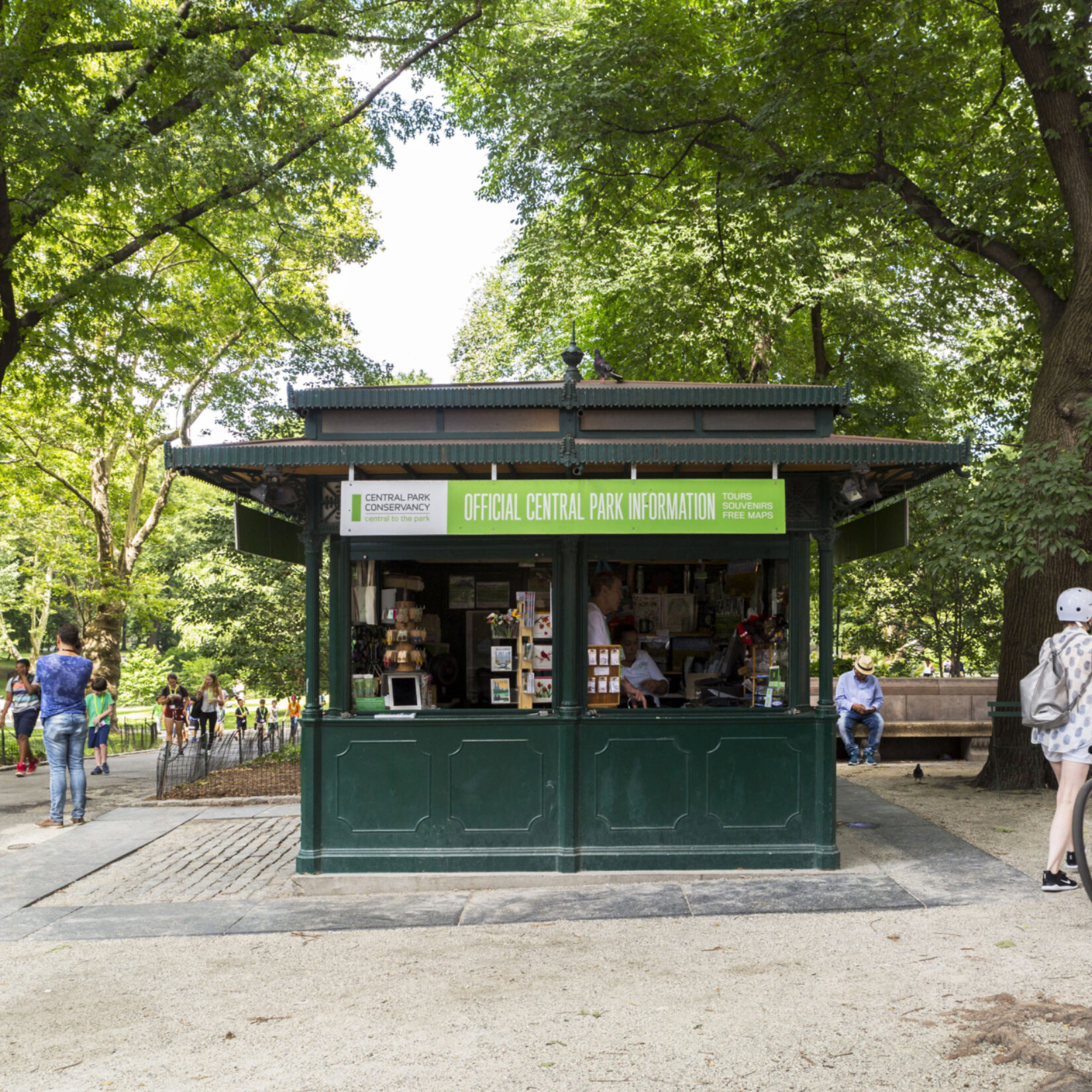 The kiosk is seen framed by summer trees and passing pedestrians.