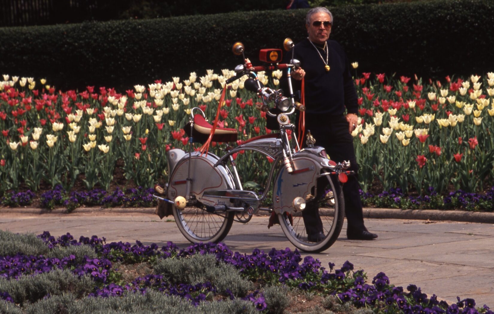 A man dressed in black walks his bicycle in the Garden. The bicycle is tricked out with a banana seat, high chrome handlebars accentuated with streamers, mirrors and an old-fashioned horn.