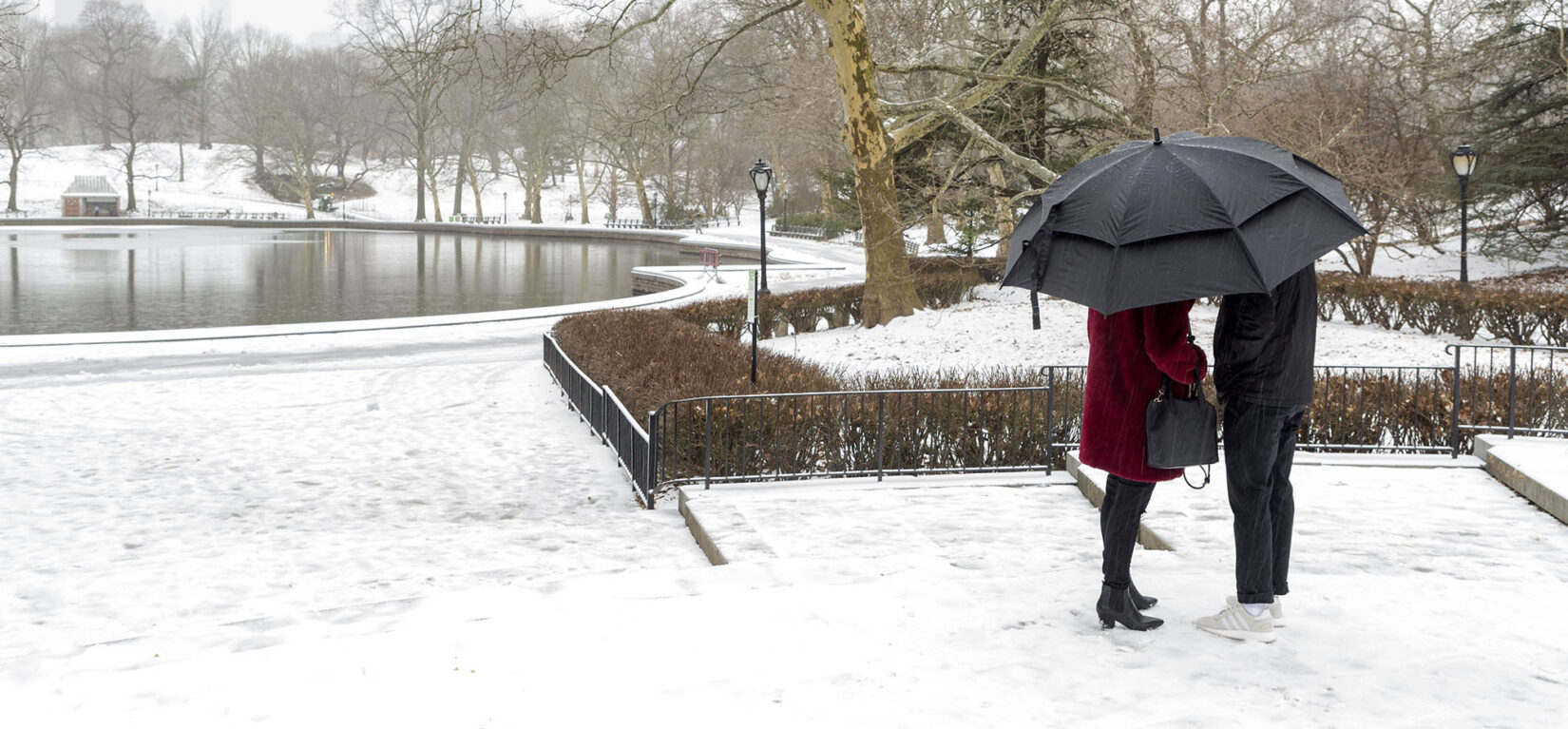 A couple huddle under an umbrella with the snow-covered Park surrounding Conservatory Water behind them