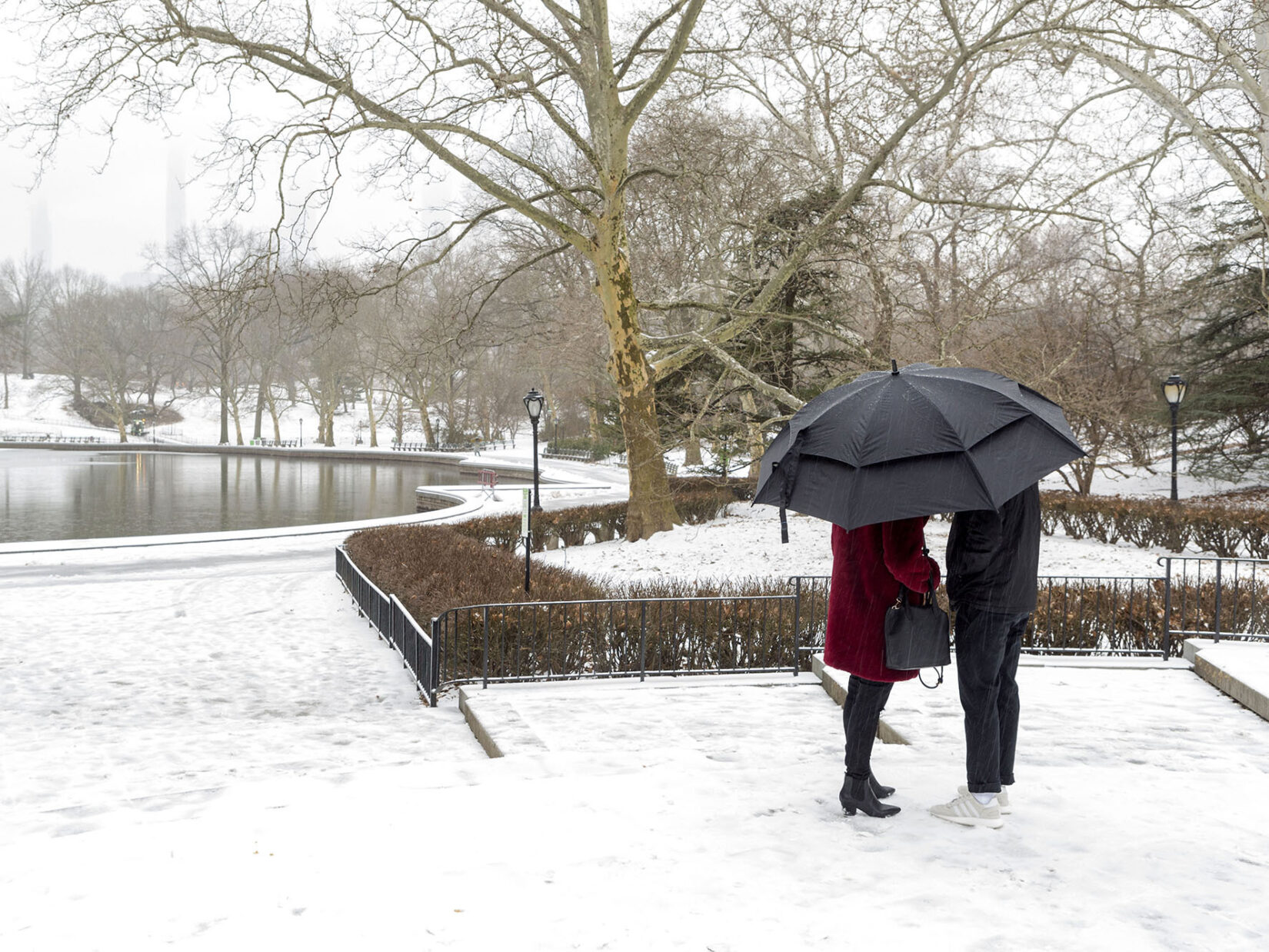 A couple huddle under an umbrella with the snow-covered Park surrounding Conservatory Water behind them