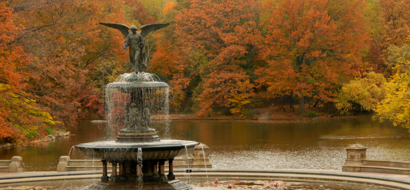 The Bethesda Fountain pictured in fall, with red and yellow foliage reflected in the Lake