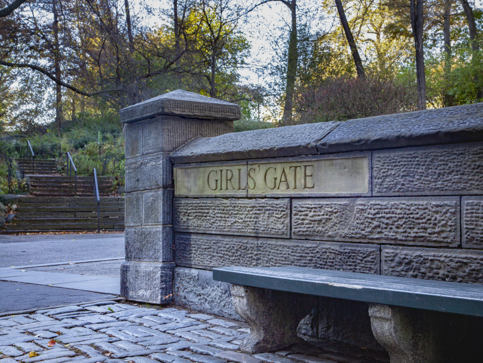 A low stone wall with "Girls' Gate" carved into it. Behind the wall is a grassy hill with many trees and bushes. In front of it is an area paved with rectangular stones and a low bench, painted green.