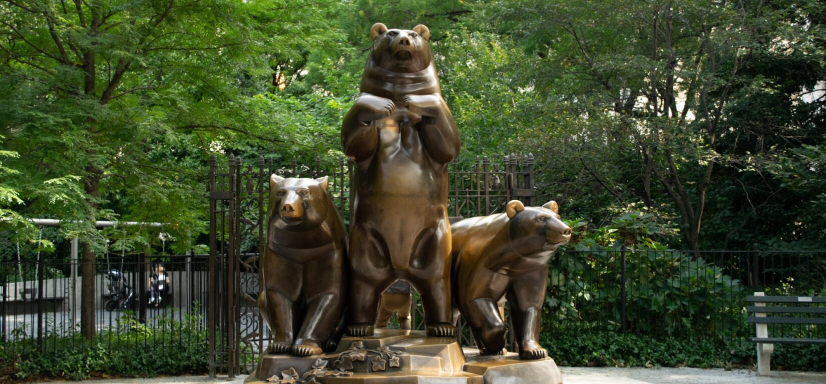 The statue standing at the center of a circular plaza, ringed by a decorative iron fence, benches, and trees