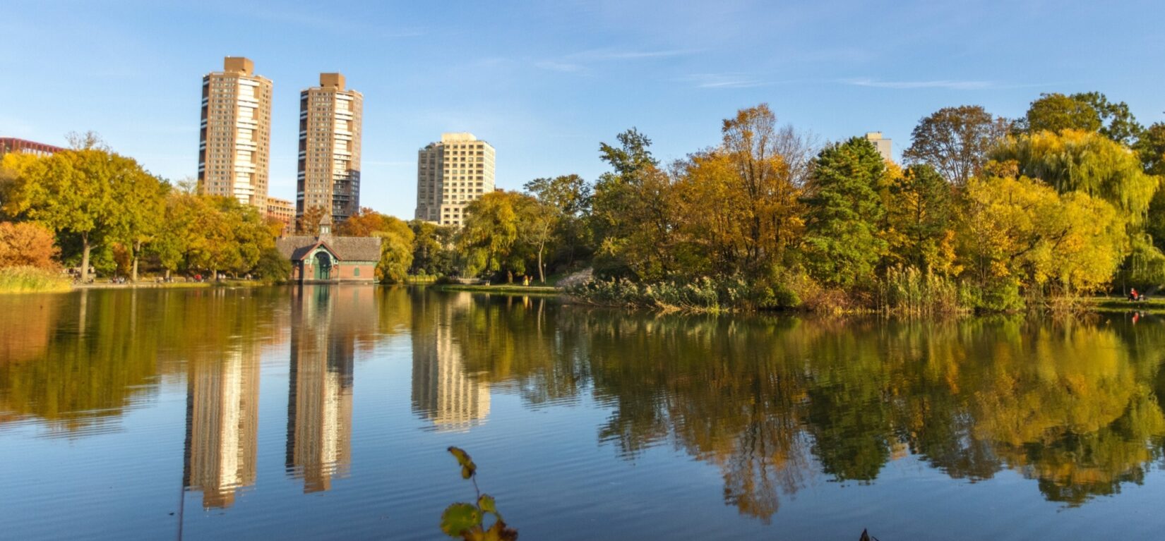 A view across the Harlem Meer, showing the Dana Discovery Center on the far shore and the skyline of Harlem behind it.