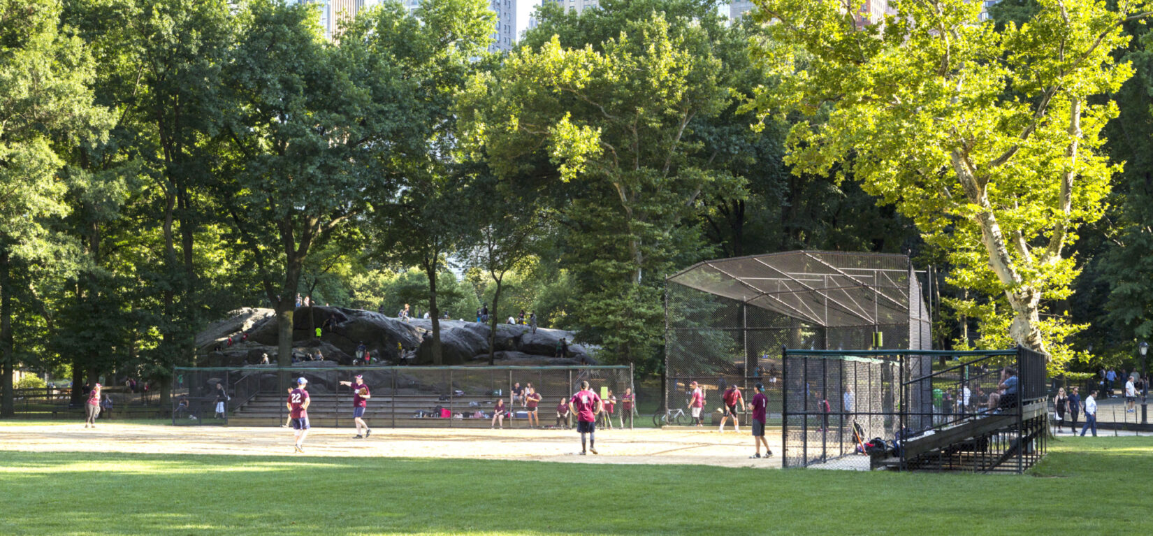 A softball game takes place under trees and the skyline of 59th Street