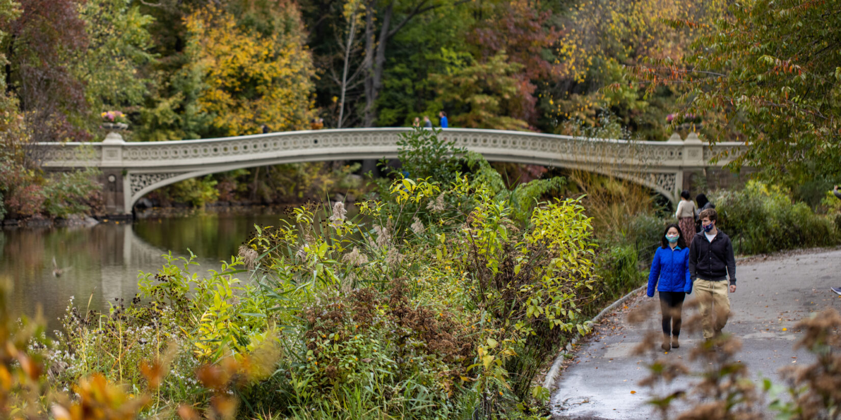 A couple walks along the Lake in autumn with Bow Bridge in the background