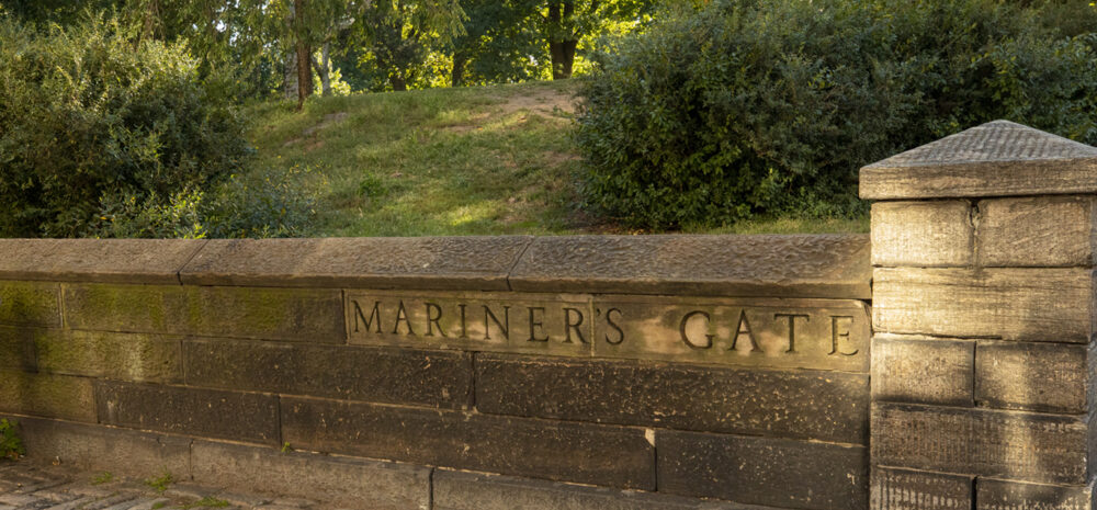 A low stone wall with the words "Mariners' Gate" carved into it. Behind the wall is a grassy hill with bushes and trees. In front of the wall the ground is paved with rectangular stones.