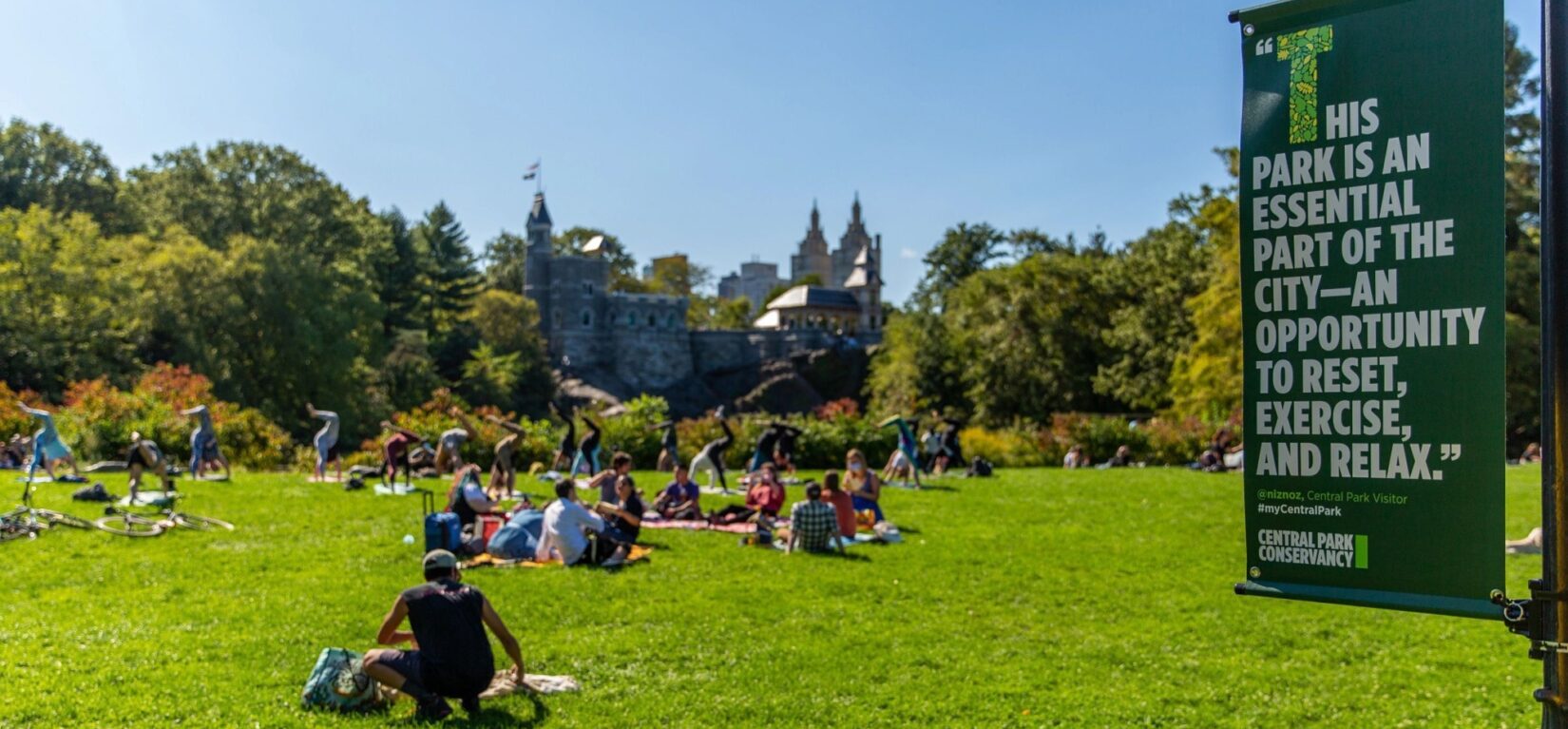 People enjoy a Park lawn under a beautiful blue sky, with a banner hanging from a lamppost entering the frame on the right. The banner has a quote about the Park.