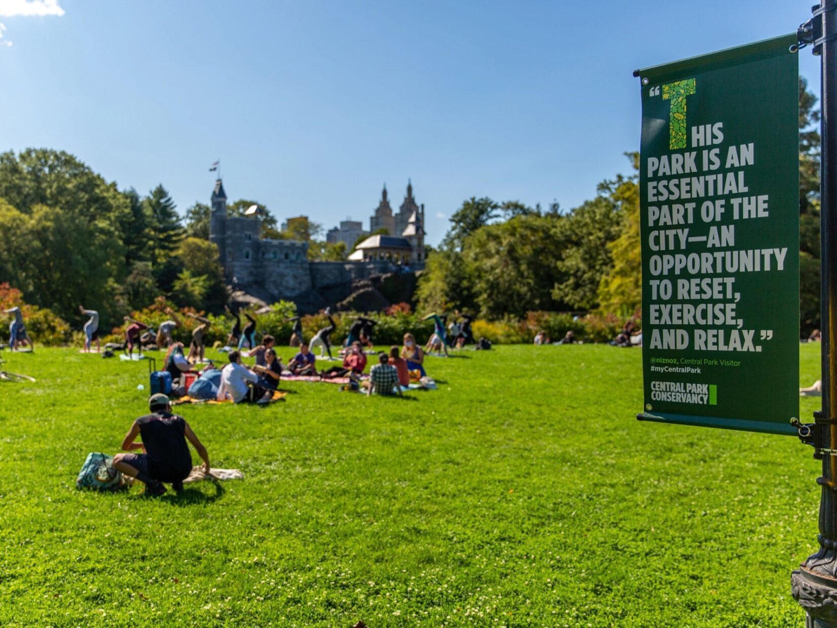 People enjoy a Park lawn under a beautiful blue sky, with a banner hanging from a lamppost entering the frame on the right. The banner has a quote about the Park.