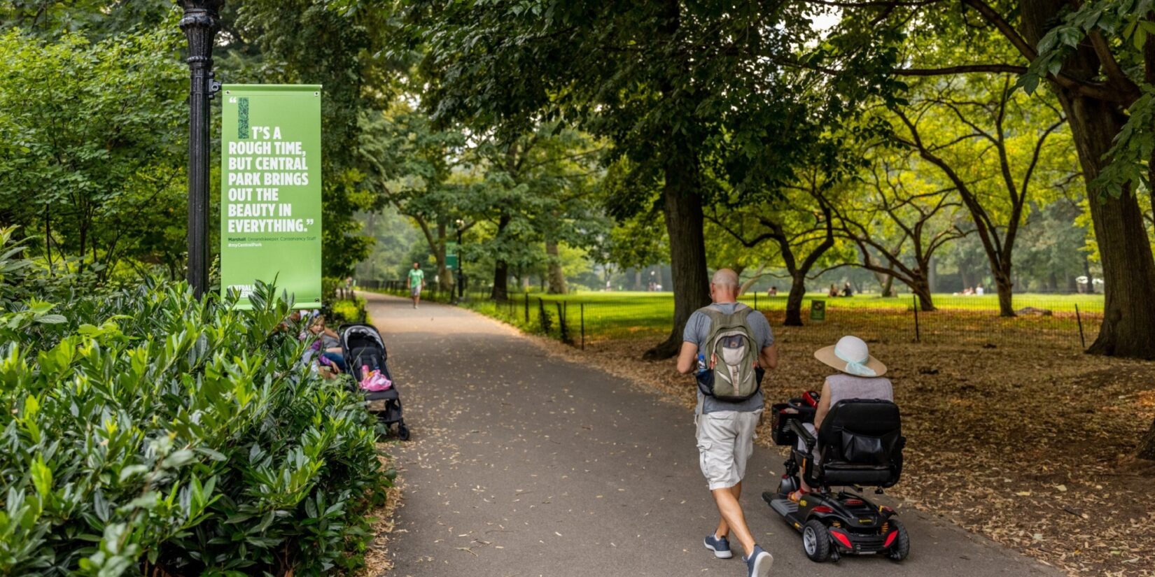 A man jogs alongside his companion, who is in a motorized wheelchair, as they pass a Conservancy banner.