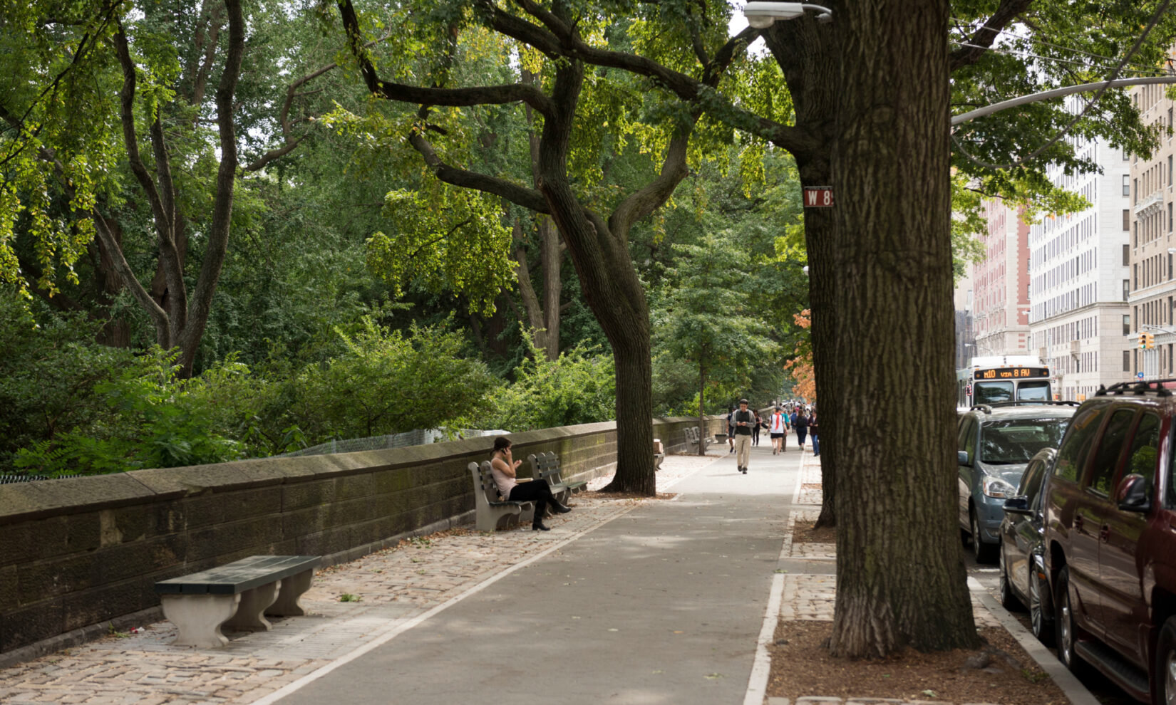 A view of the perimeter in summer, with pedestrians in the distance, one of them sitting on a park bench.