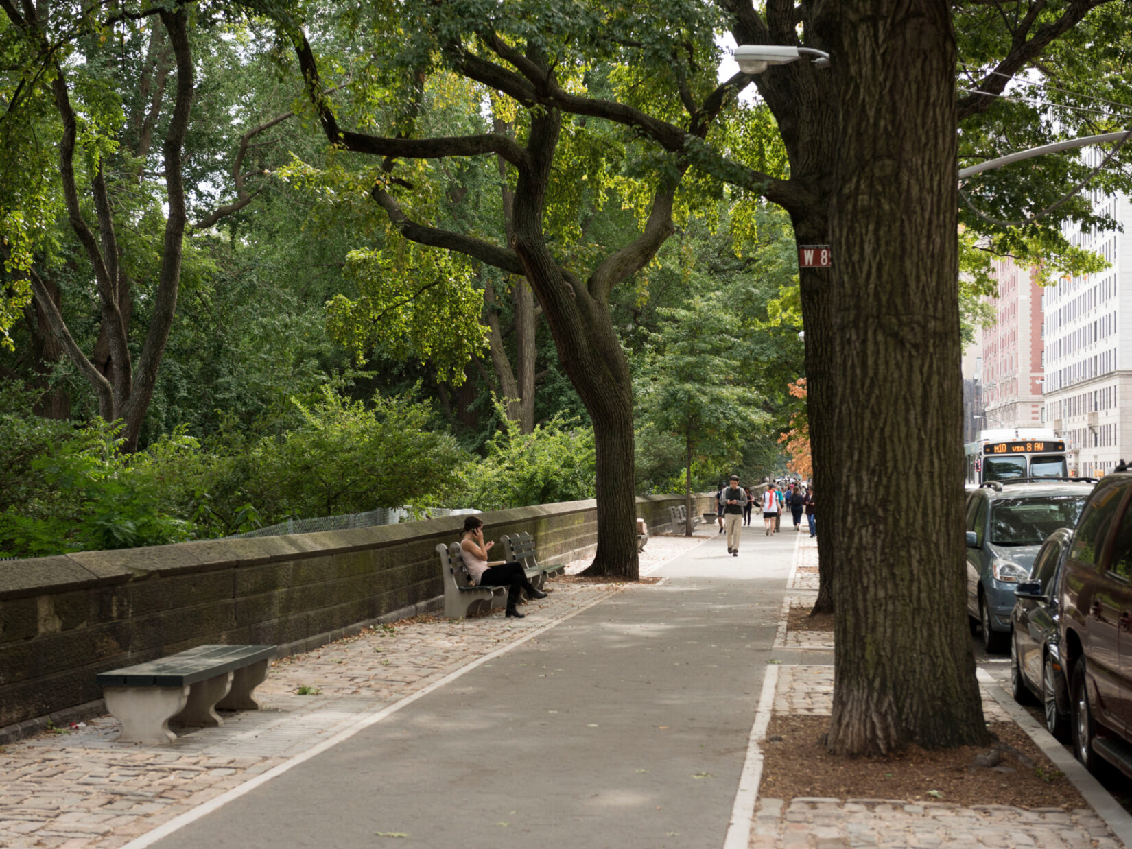 A view of the perimeter in summer, with pedestrians in the distance, one of them sitting on a park bench.