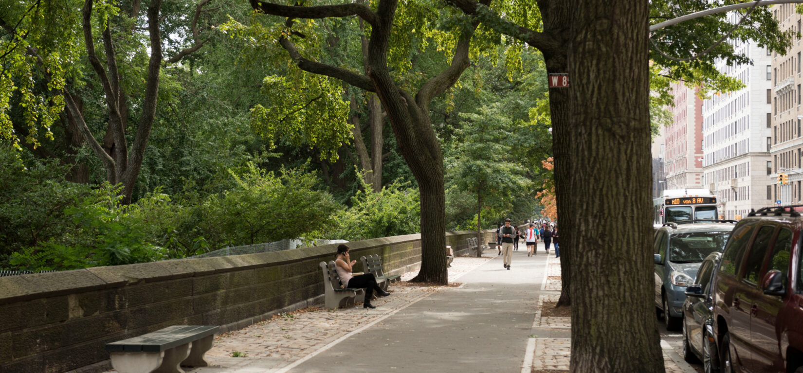 A view of the perimeter in summer, with pedestrians in the distance, one of them sitting on a park bench.