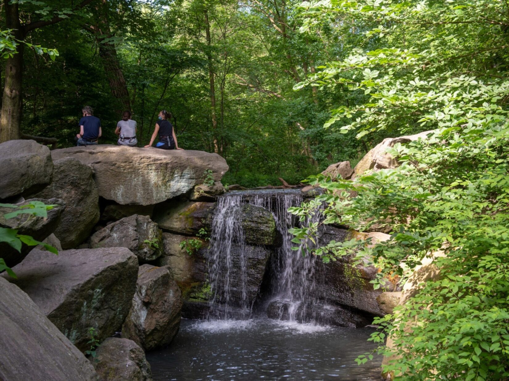 Three park-goers sit on a rock beside a waterfall overlooking the Ravine