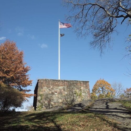 The American flag waves at the top of a pole mounted on the Blockhouse, against a clear blue sky.