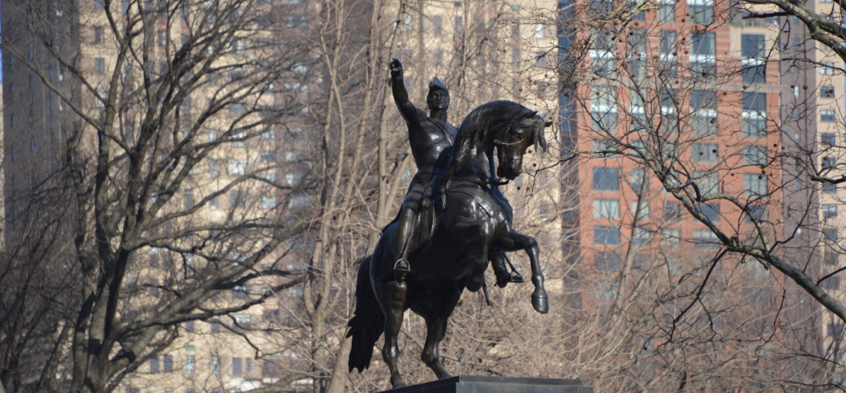 The equestrian statue shown in winter, with buildings shown through bare branches in the background