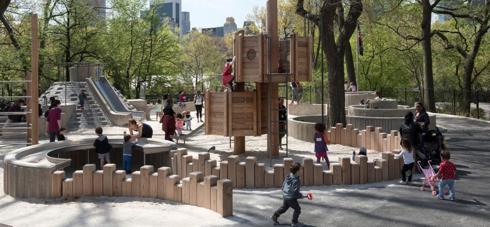 Children play late on an early spring day in a sand area ringed by vertical wooden beams of random heights.