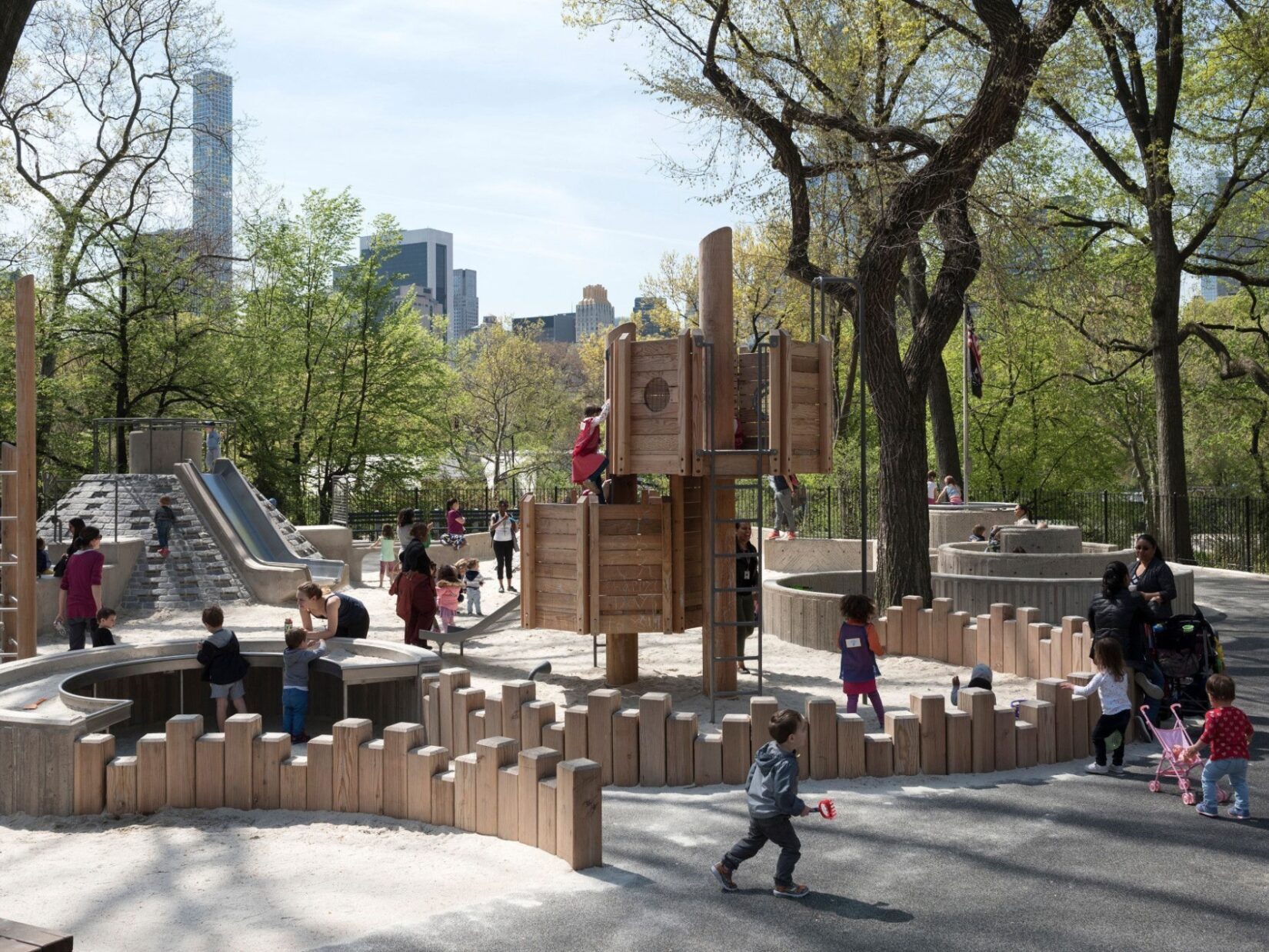 Children play late on an early spring day in a sand area ringed by vertical wooden beams of random heights.