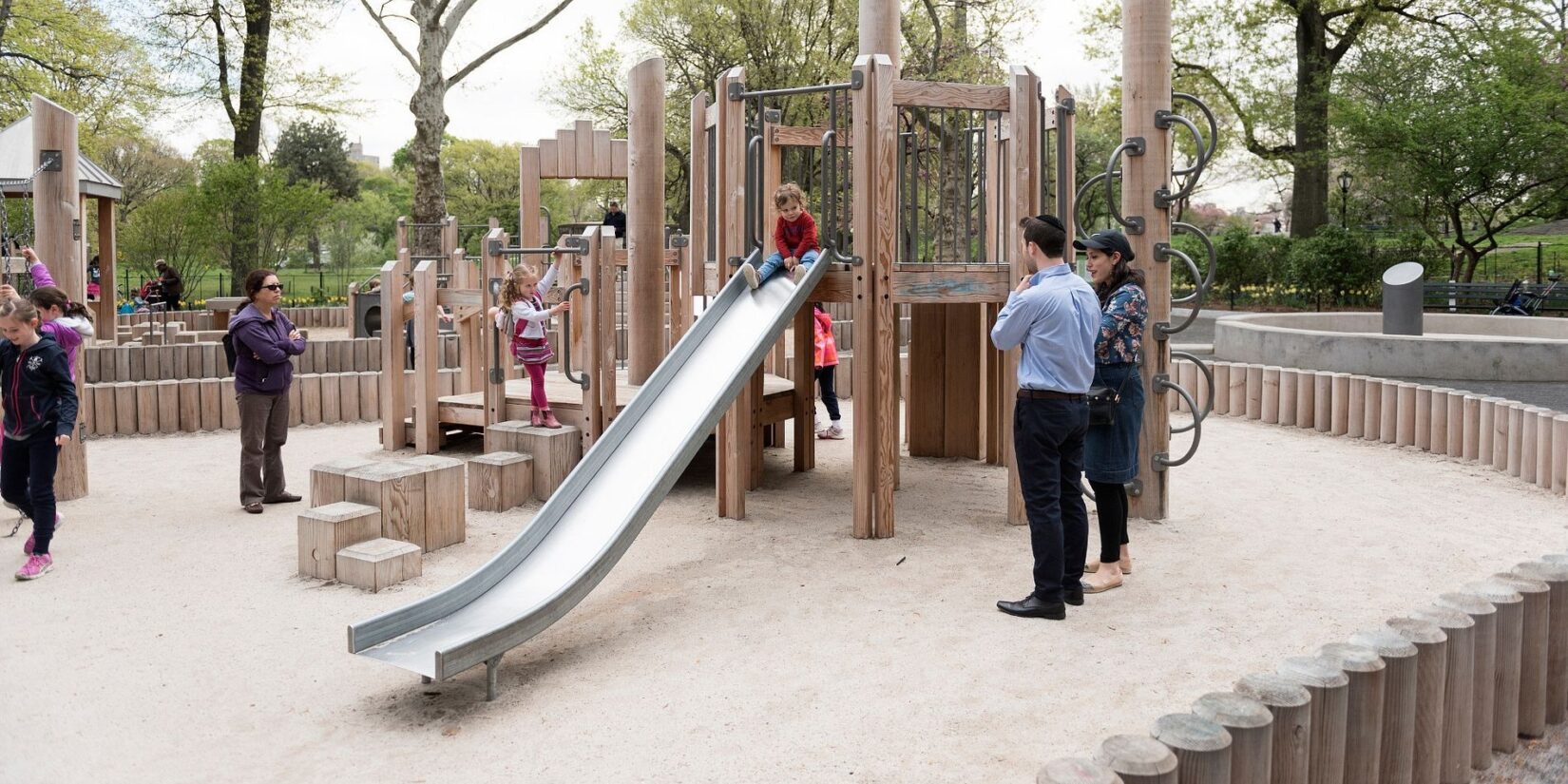 A few parents watch as a toddler prepares to slide down the sliding pond