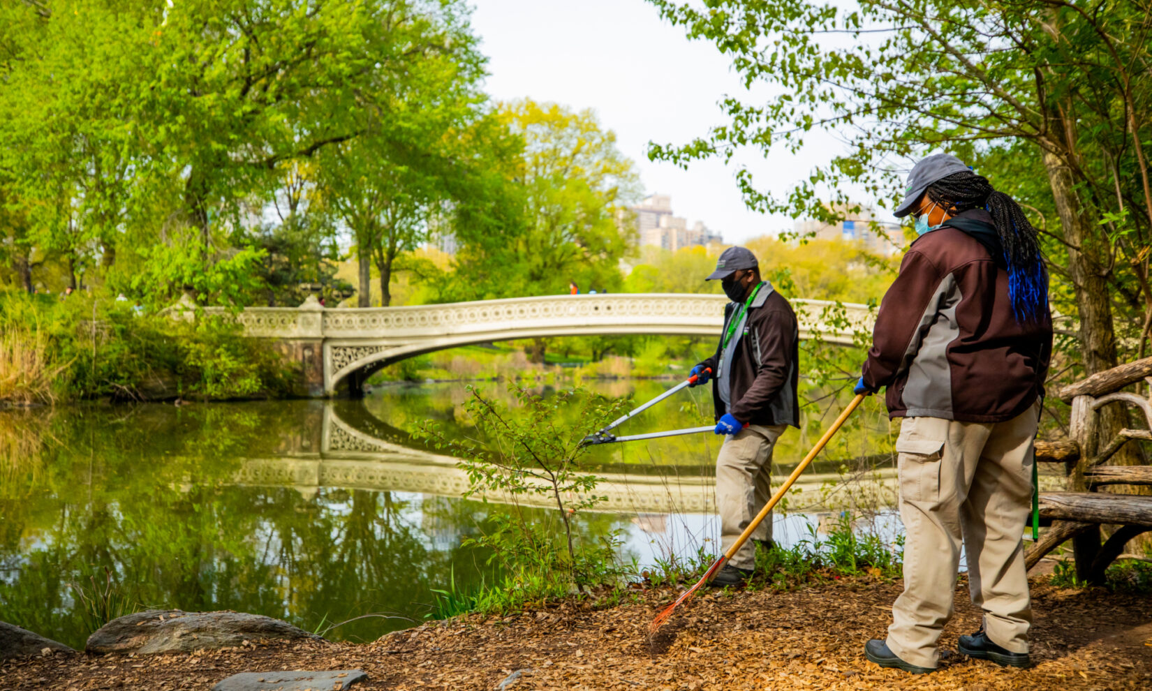 Two staff members tending to the embankment of the Lake with Bow Bridge in the background