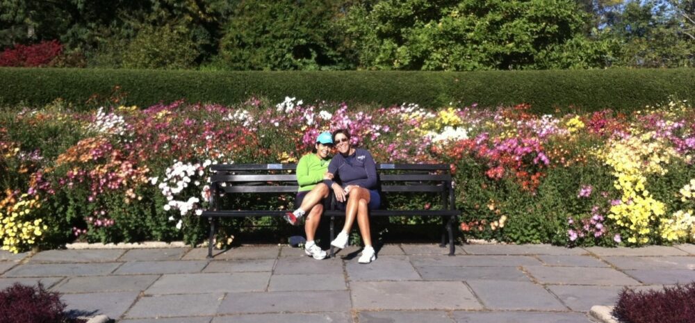 Martha and Serena sitting on a Park bench in front of colorful flowers.