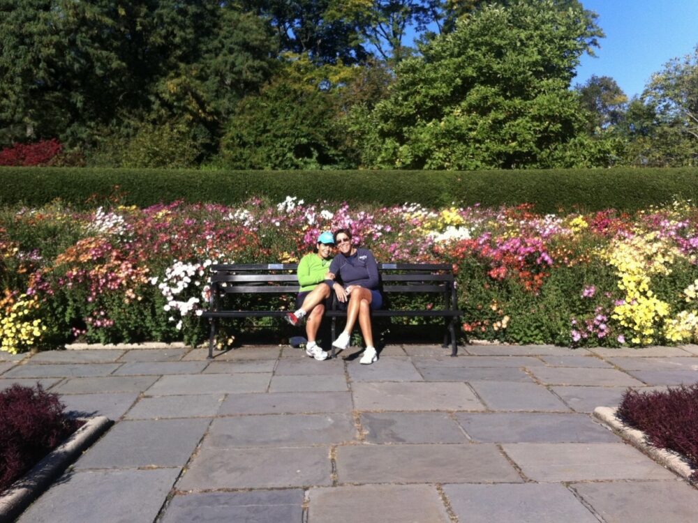 Martha and Serena sitting on a Park bench in front of colorful flowers.