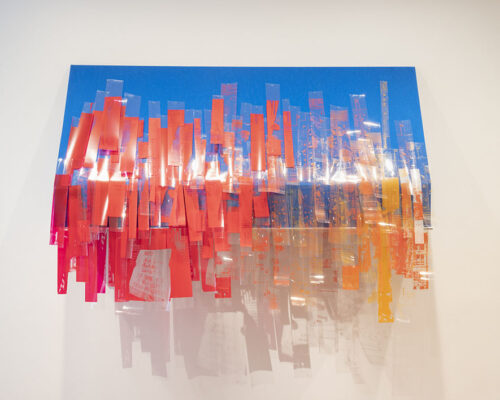 A sculptural piece photographed in a galler, featuring strips of red and transparent plastic drooping from a blue rectangle