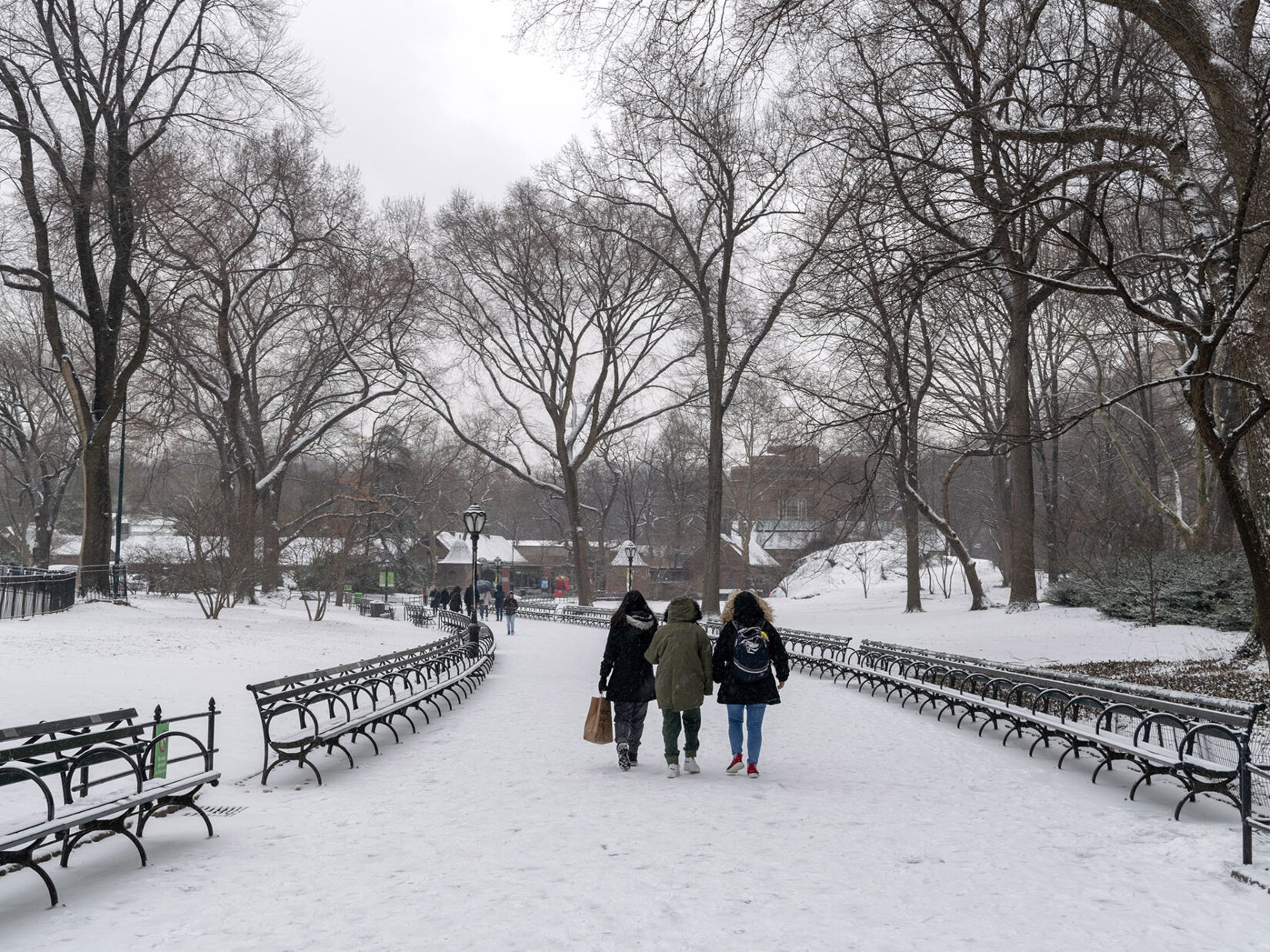 Three parkgoers bundled up for winter walk a snowcovered, bench-lined path in the Park