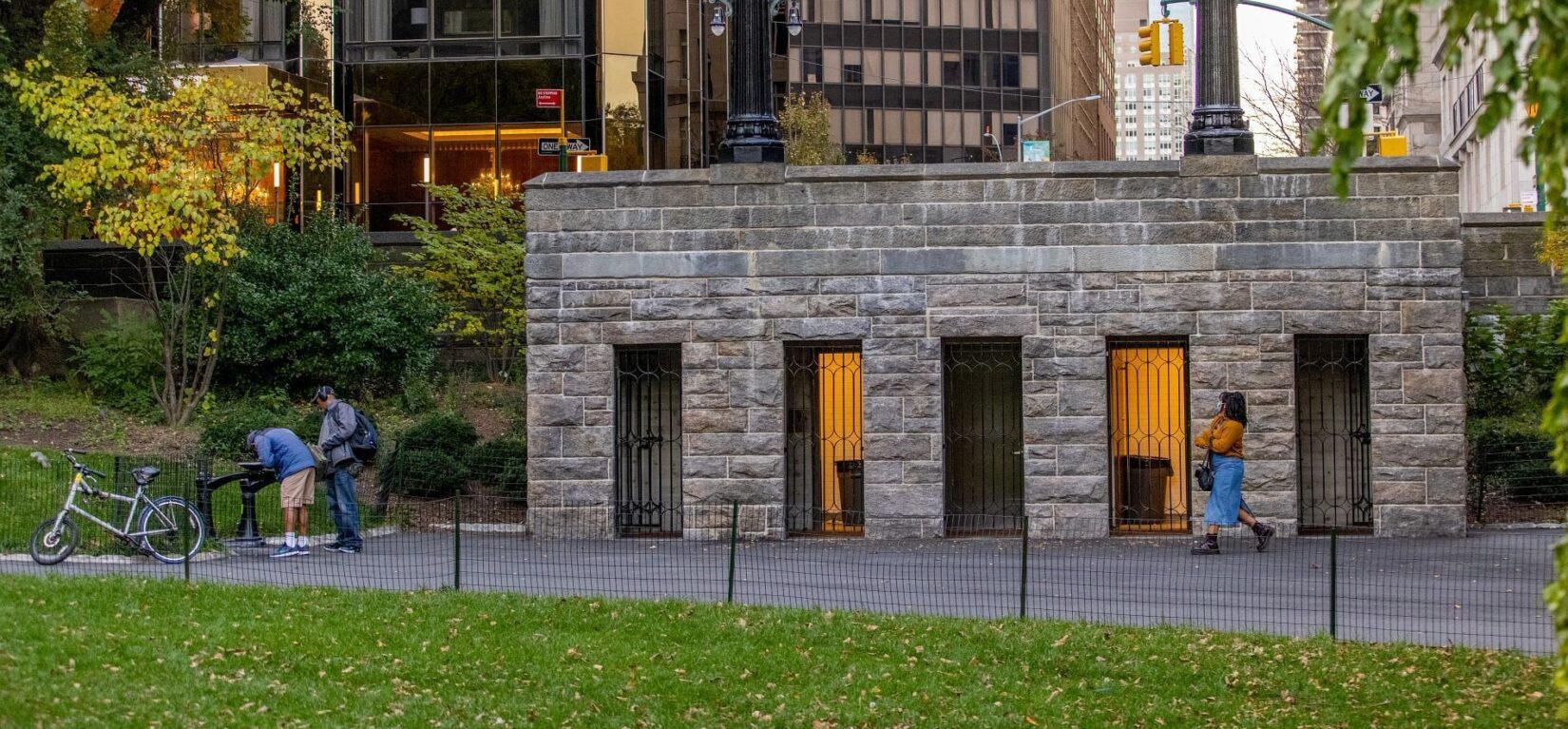 The public restroom seen in early twilight, nestled in the landscape of the Park with the buildings of on the other side of Central Park West in the background.