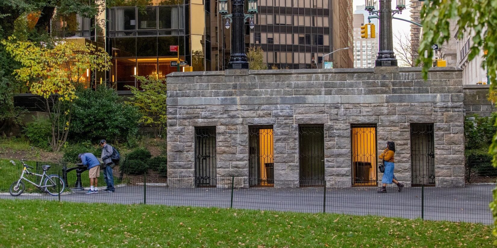 The public restroom seen in early twilight, nestled in the landscape of the Park with the buildings of on the other side of Central Park West in the background.