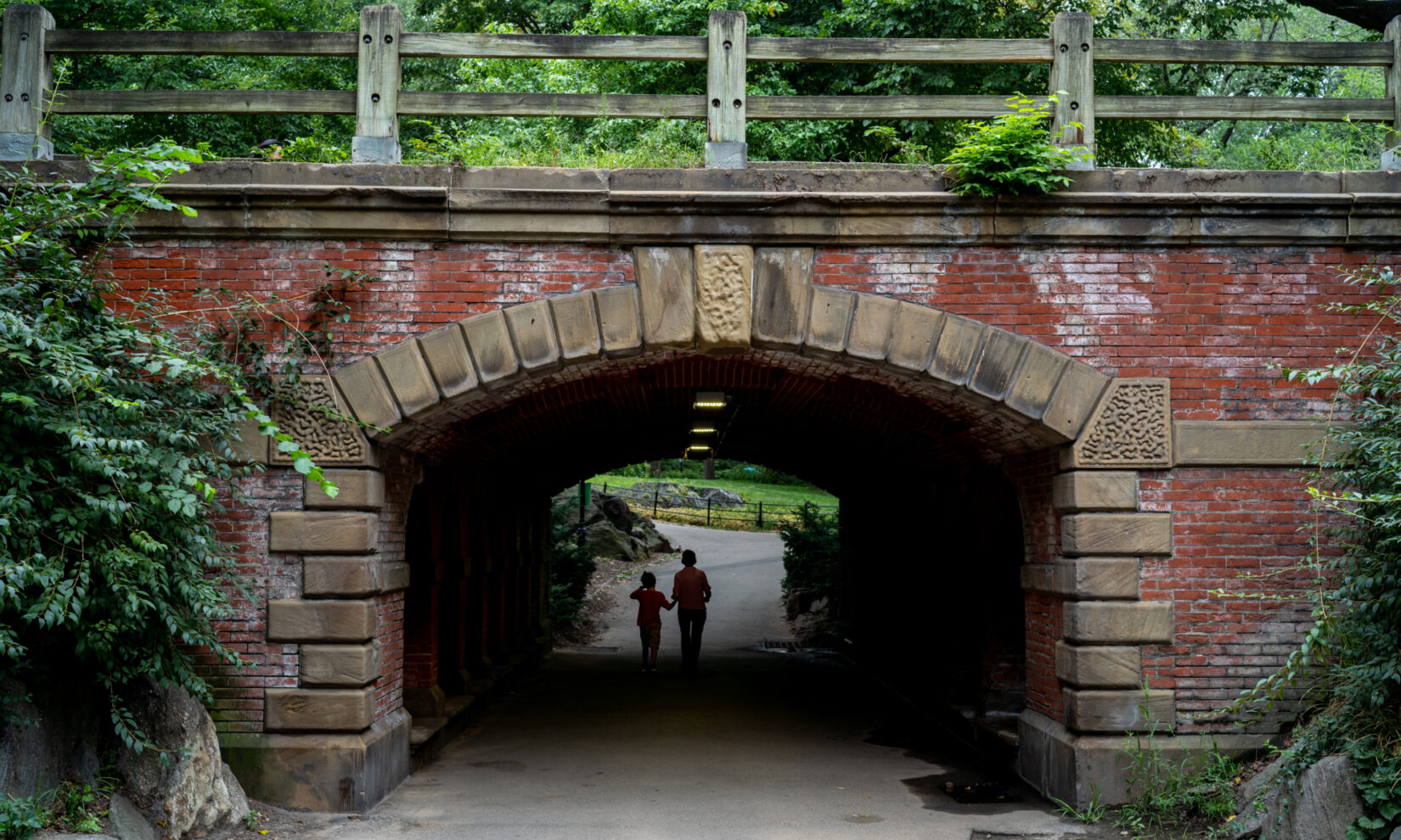 Two park-goers are silhouetted in the shadow under the arch and the bright light of a summer day