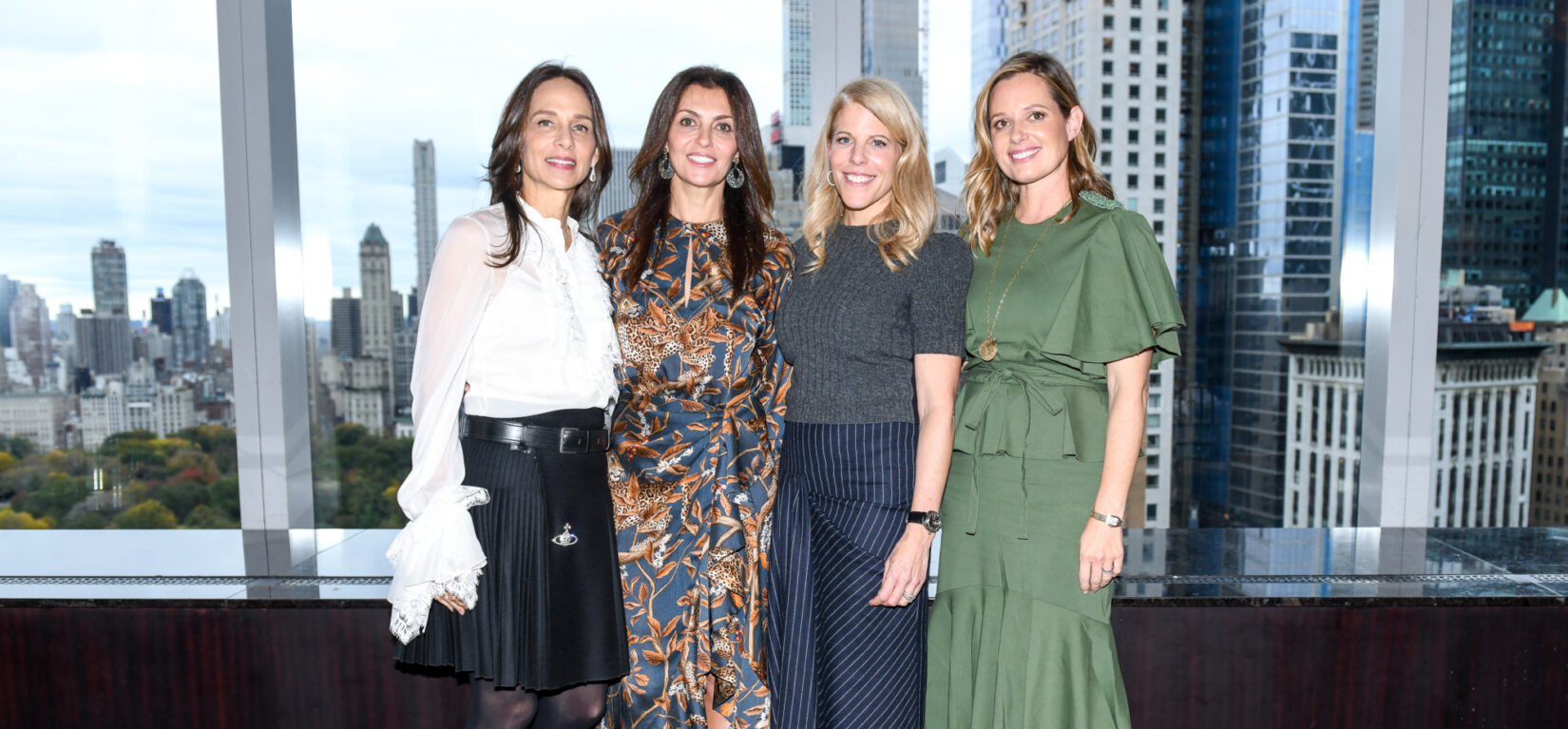 Members of the Women's Committee pose in front of a window with the NYC skyline behind them