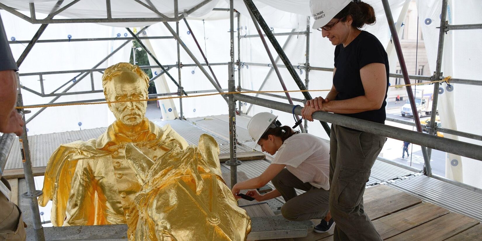 The top of the sculpture is seen from within its surrounding scaffolding, being worked on by two artisans.